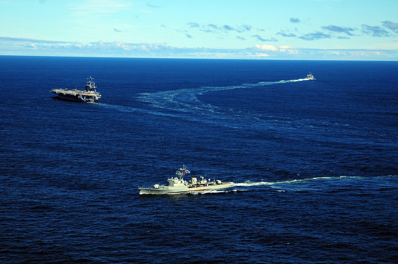 A grey Uruguayan navy frigate sails in the Atlantic with a US aircraft carrier and cruiser in the distance