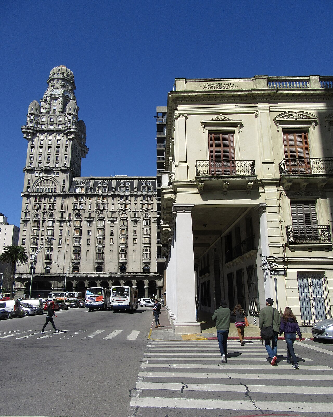 Pedestrians cross a street beside Plaza Independencia in central Montevideo, with the Palacio Salvo tower and city buses in the background