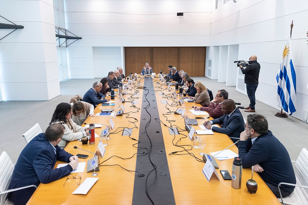 Uruguay's cabinet ministers seated along a long table in a meeting room, with the blue and white striped Uruguayan flag at right and a cameraman standing by