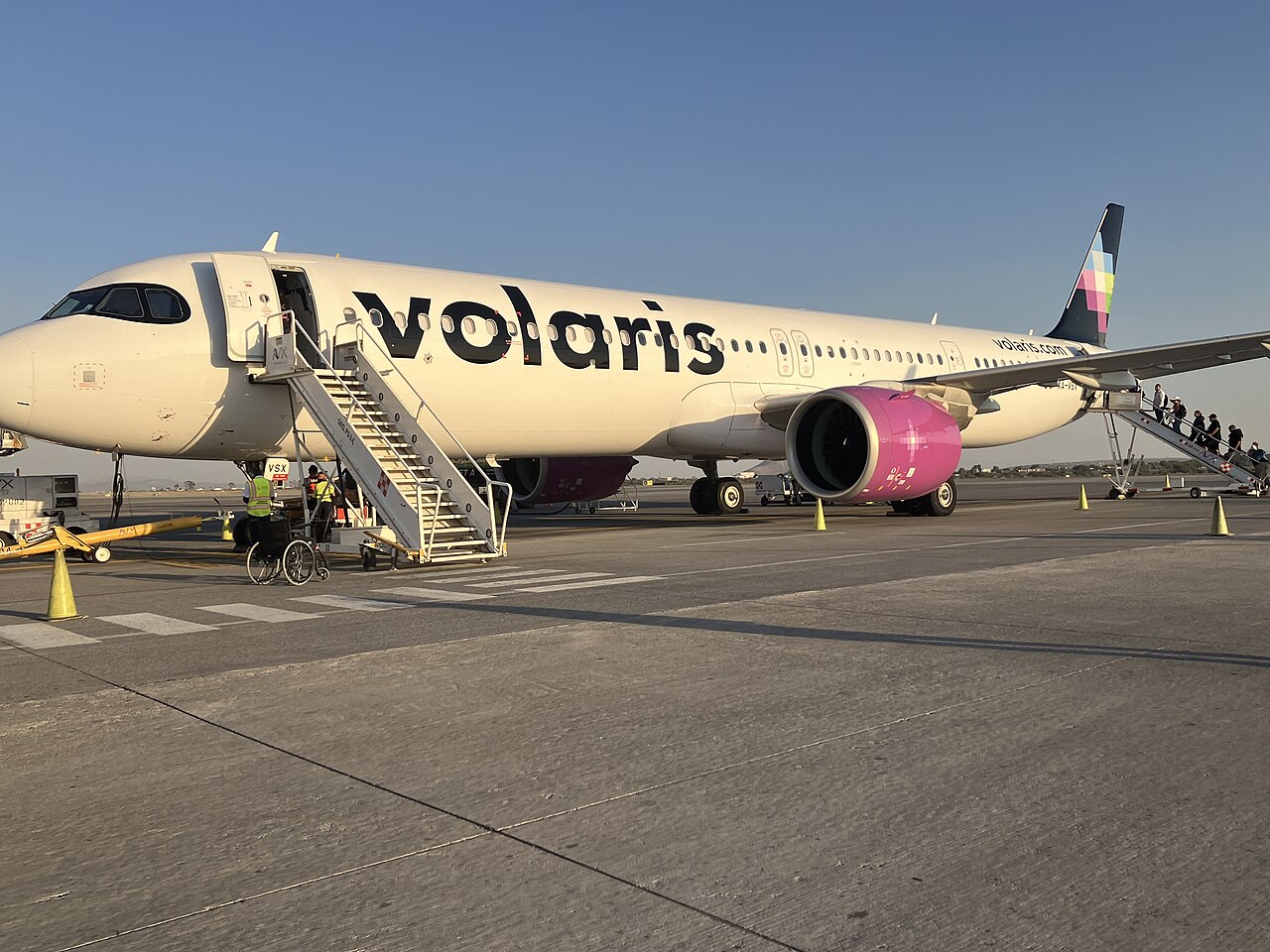 Passengers board a white Volaris Airbus A321neo with pink engines by stairs at the front and rear doors on a sunny apron