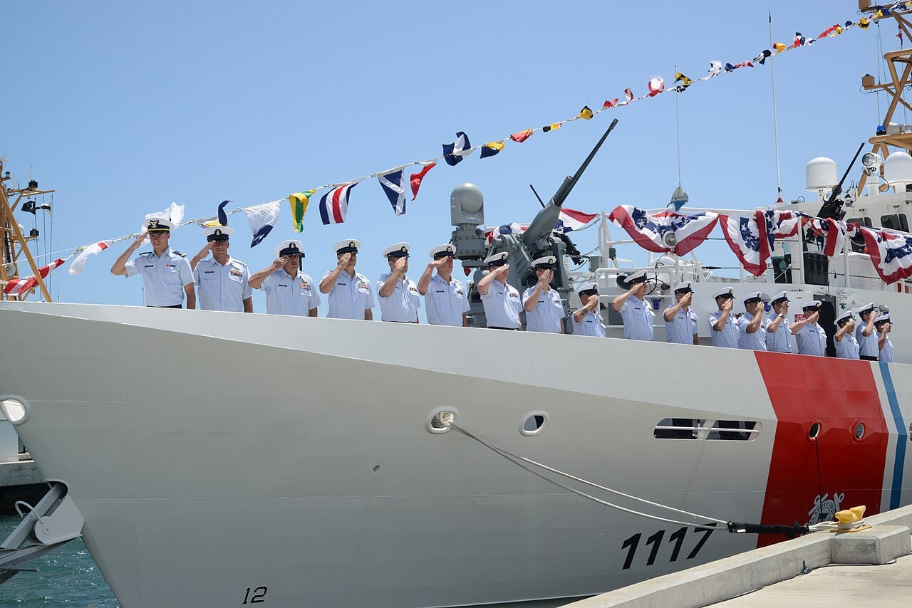 Coast Guard crew in light-blue uniforms saluting on the deck of fast response cutter 1117, decorated with signal flags and bunting at a pier