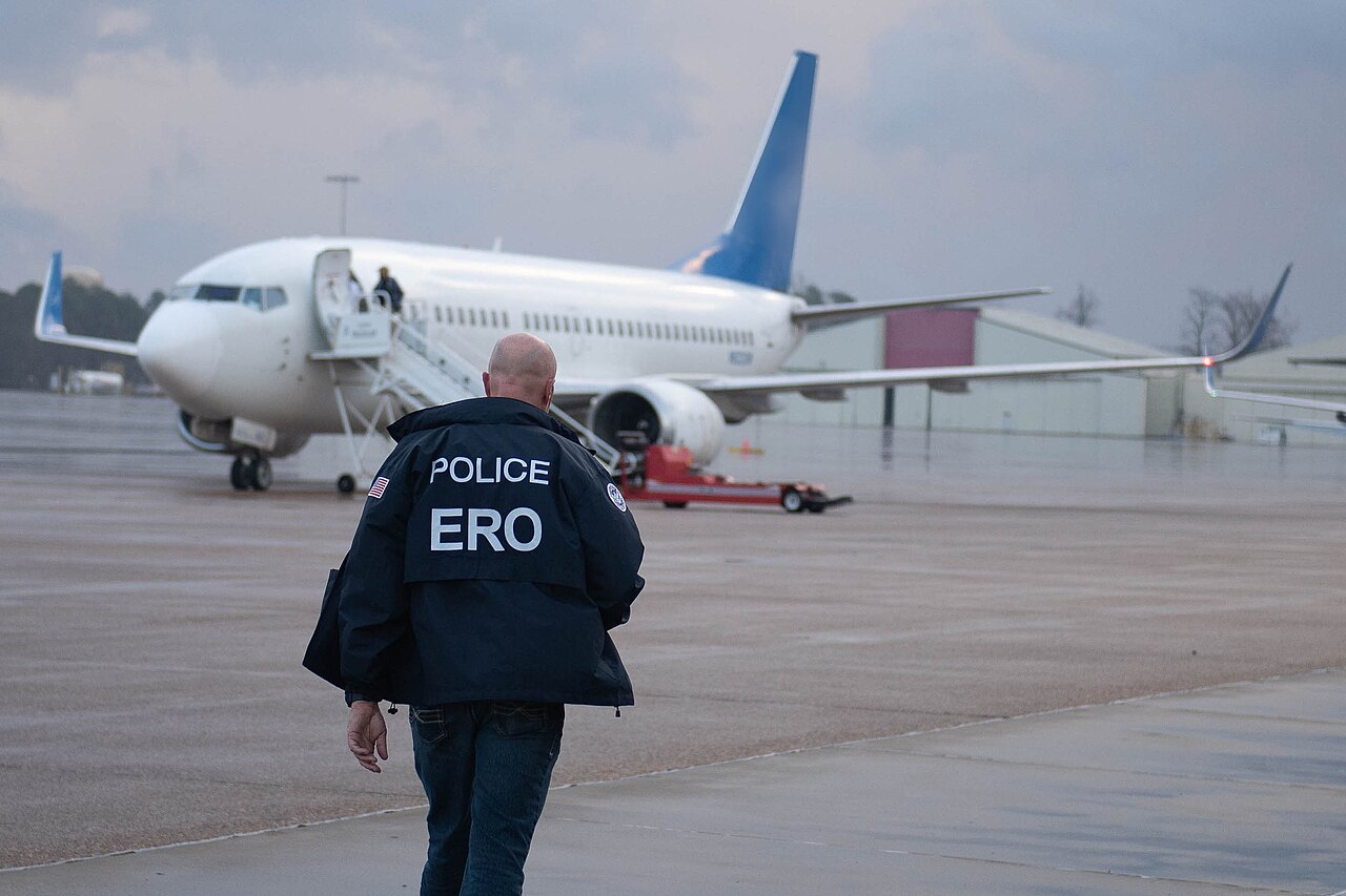 A US immigration officer in a dark jacket marked Police ERO walks toward a white charter jet parked on a wet airport apron