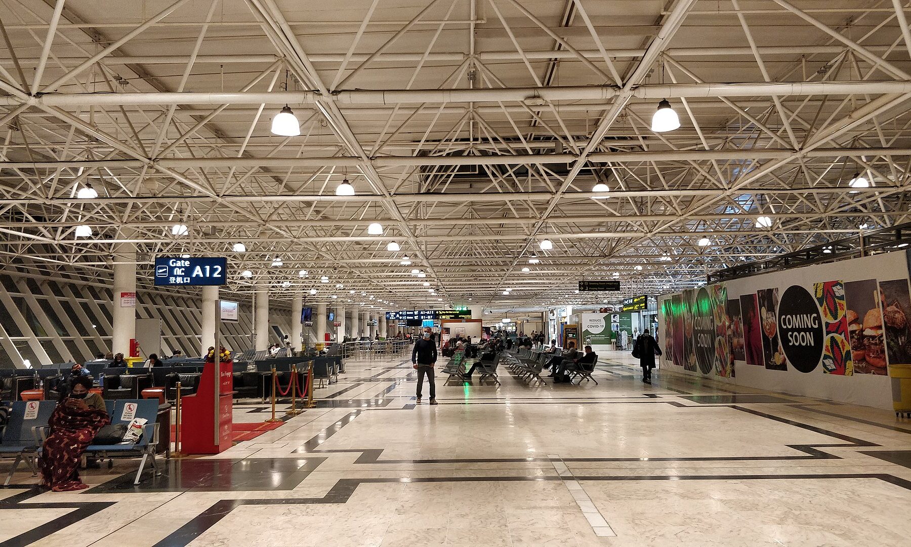 A long departures hall with white steel roof trusses and travellers seated near Gate A12 at Bole airport in Addis Ababa