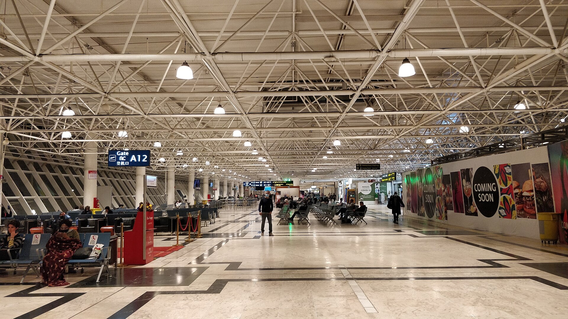 A long departures hall with white steel roof trusses and travellers seated near Gate A12 at Bole airport in Addis Ababa