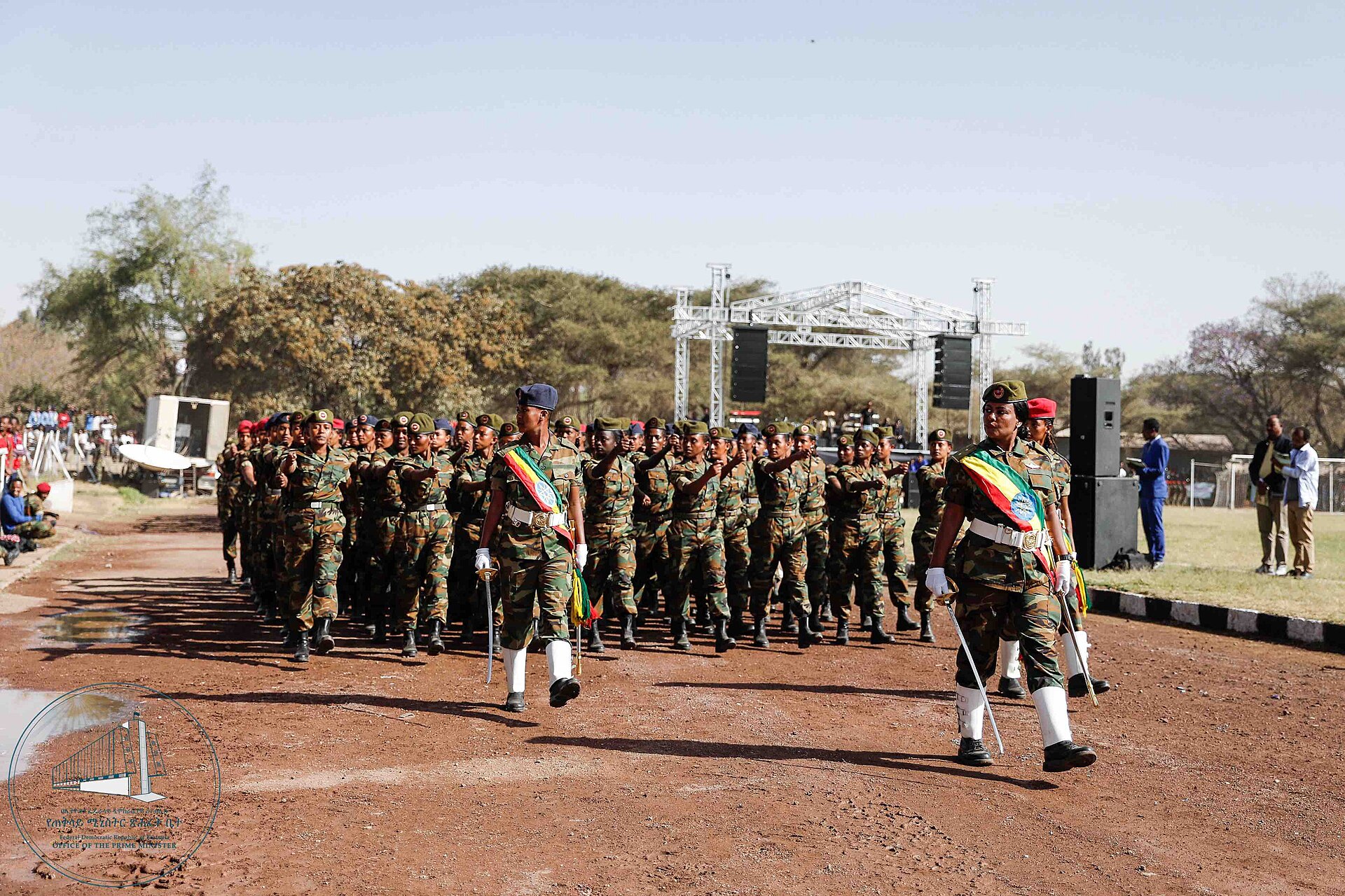 Rows of Ethiopian soldiers in camouflage uniforms march on a red dirt road past a stage, led by two officers wearing sashes in the national colours