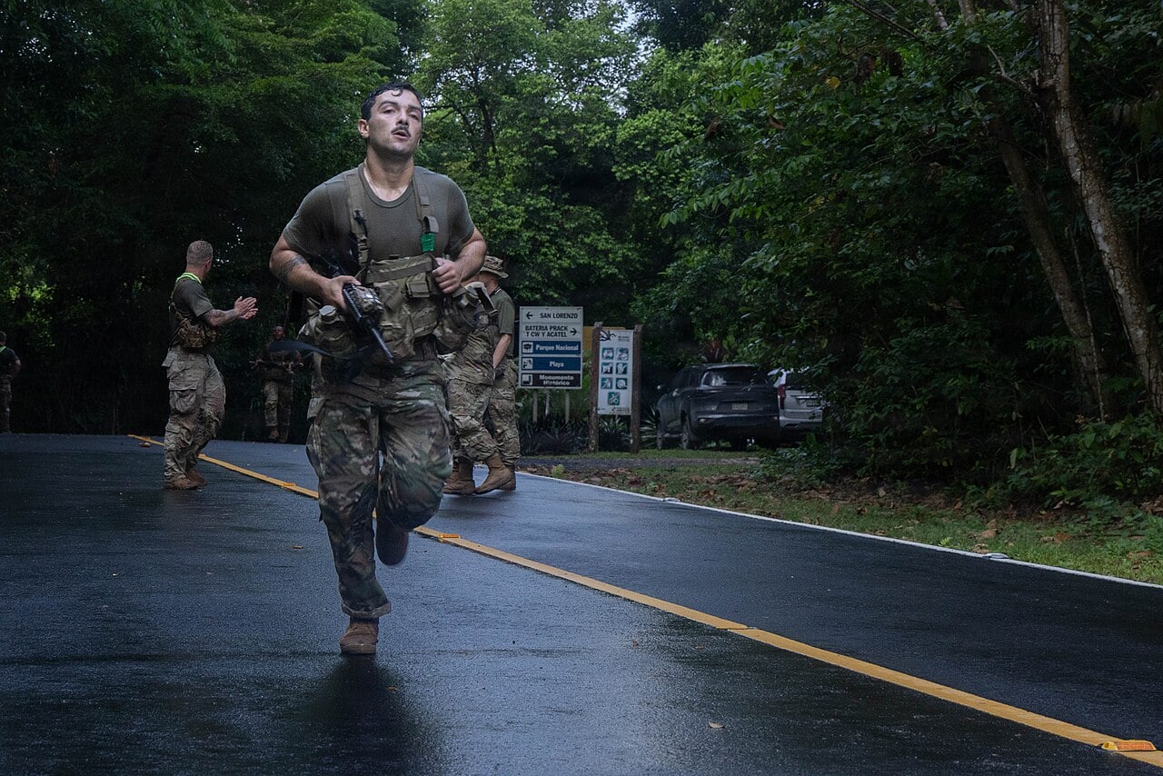 A US soldier in camouflage carrying a rifle runs along a wet jungle road past a sign for San Lorenzo, with other soldiers behind him