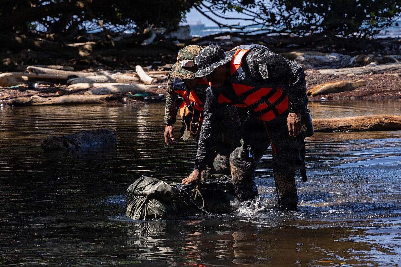 Two Panamanian security personnel in orange life vests drag a waterproof pack through shallow water near a rocky beach