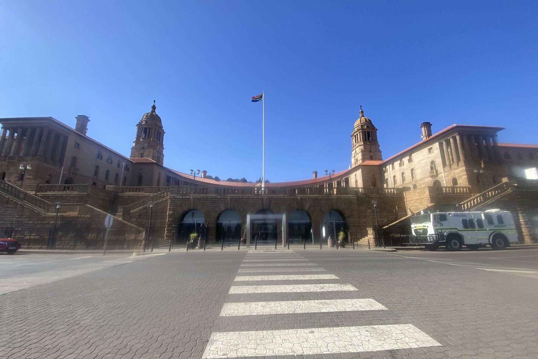 Sandstone Union Buildings with two domed towers and a flagpole flying the South African flag under a clear blue sky, a police vehicle parked at right