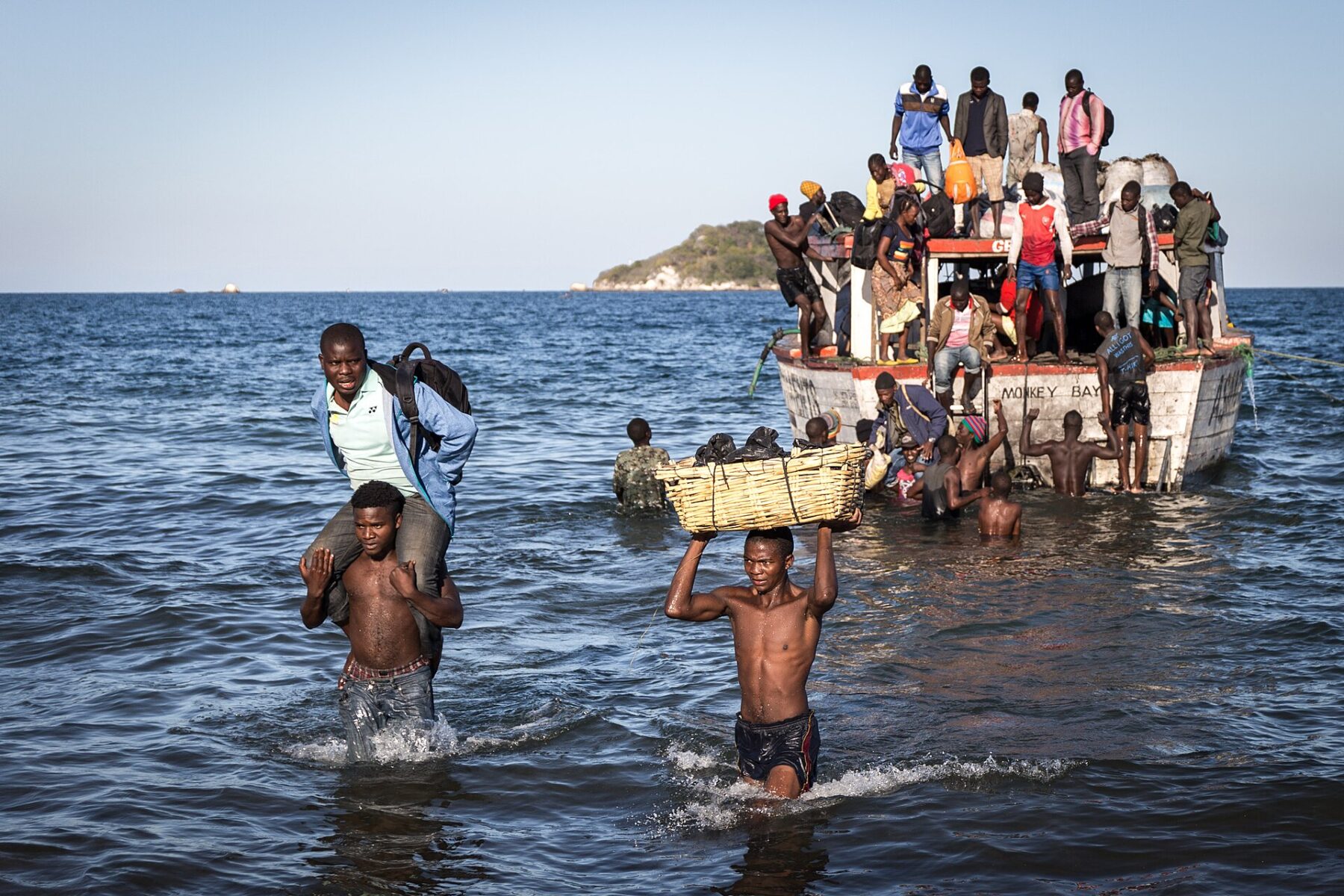 Passengers wade ashore from a crowded wooden boat on Lake Malawi while men carry a passenger and a basket through the water