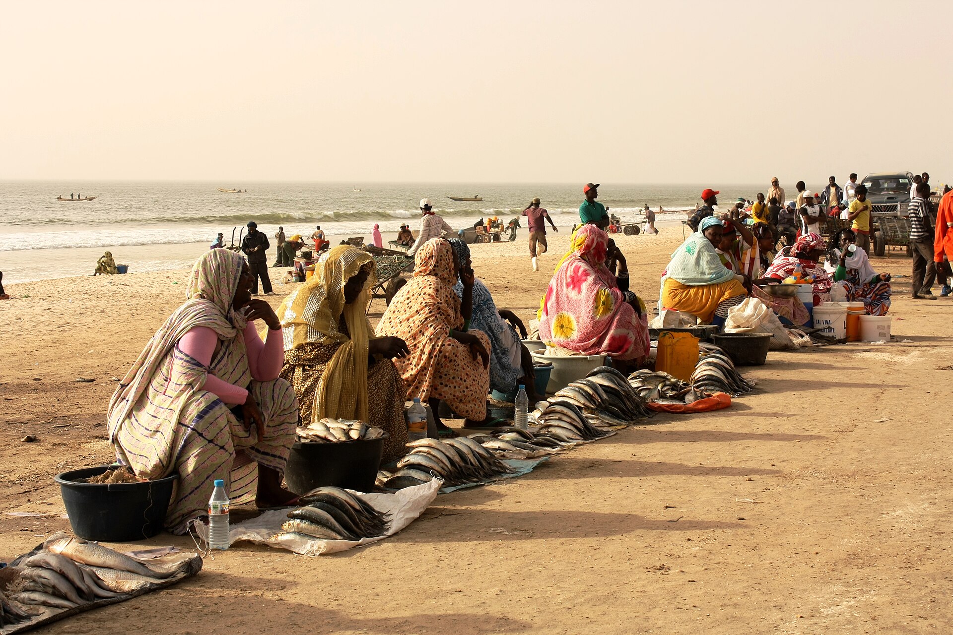 Women in colourful melhfa wraps sit on the sand beside rows of fresh fish, with fishing boats on the sea behind