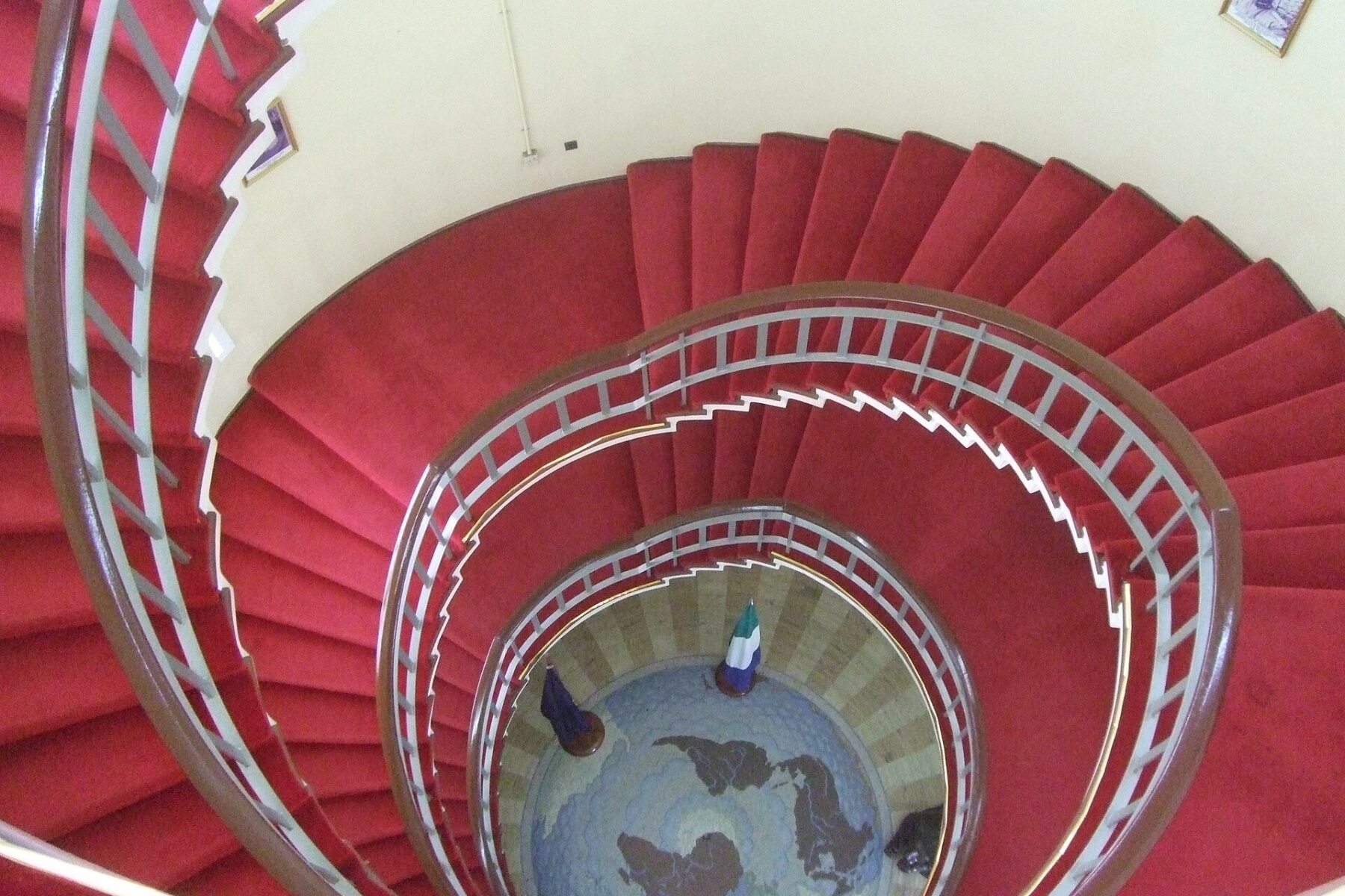 Red-carpeted spiral staircase seen from above inside State House in Freetown, with a Sierra Leone flag on the floor below