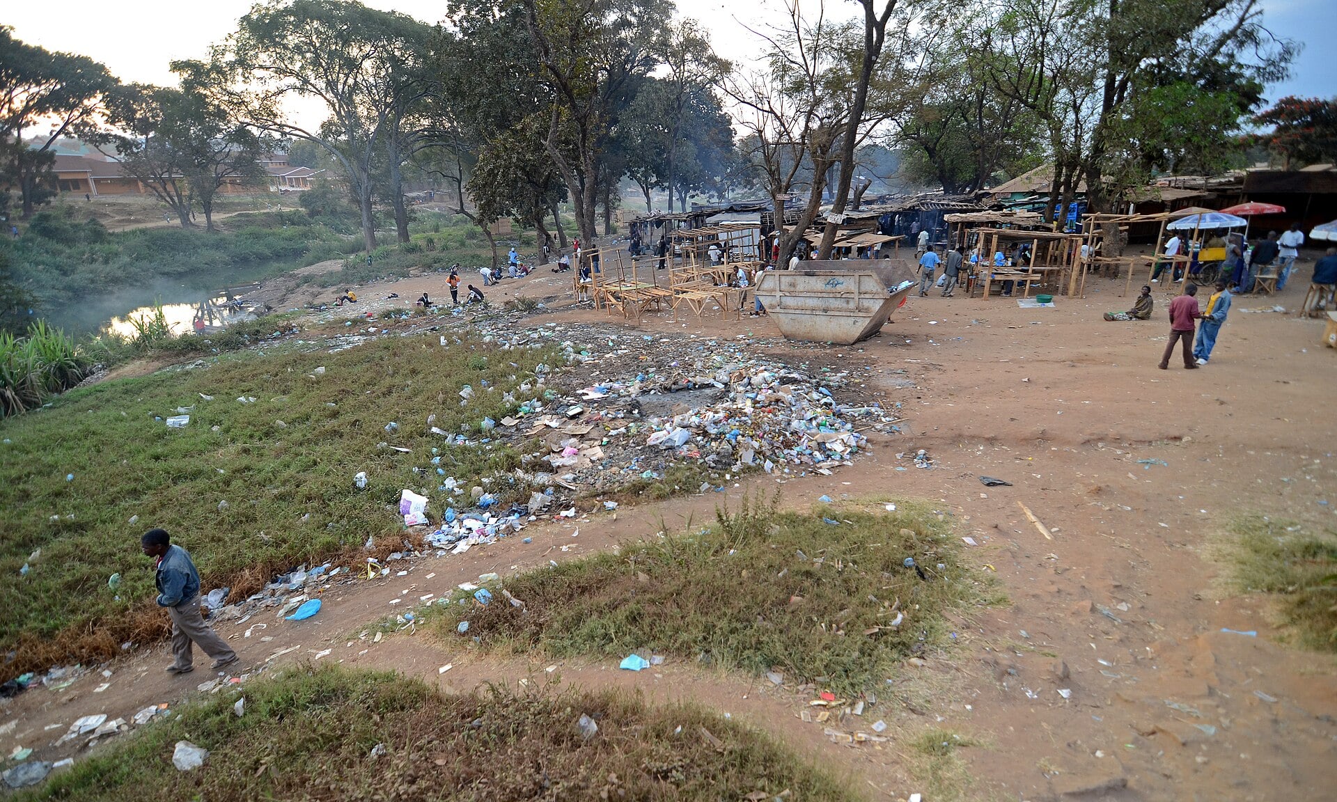 Wooden market stalls under trees beside a river in Lilongwe, with litter on the ground in the foreground and people walking on a dirt path