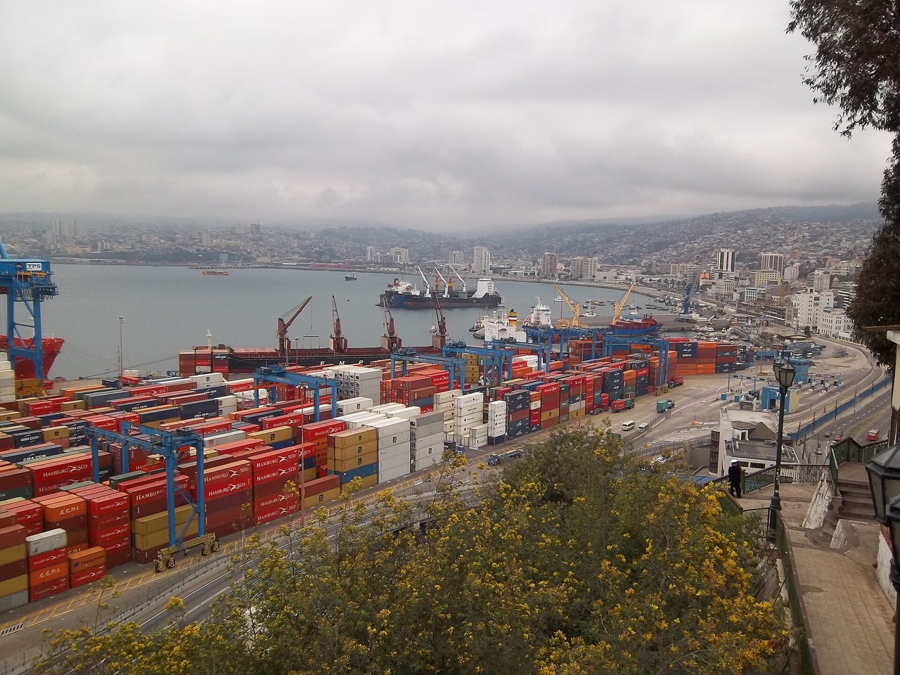 Container stacks, blue gantry cranes and ships at the port of Valparaíso, Chile, seen from a hillside with the city along the bay