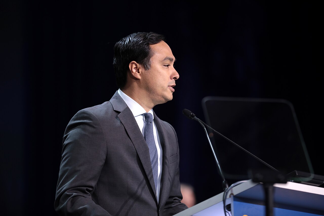 US Congressman Joaquin Castro in a grey suit and blue tie speaking at a lectern with a microphone, against a dark background