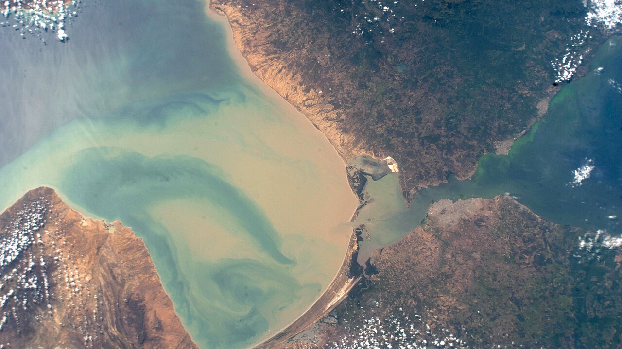 Astronaut photograph of the Gulf of Venezuela, with swirls of sediment in turquoise water, brown coastline and the narrow channel towards Lake Maracaibo