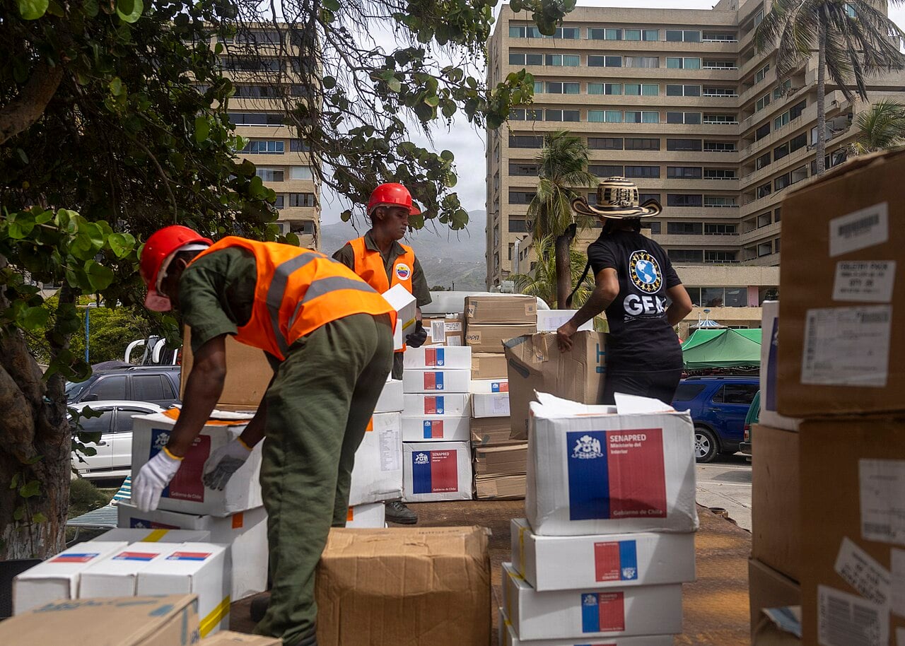 Two Venezuelan soldiers in orange vests and red helmets and a woman in a black GEM T-shirt and wide-brimmed hat stack cardboard aid boxes beside high-rise buildings