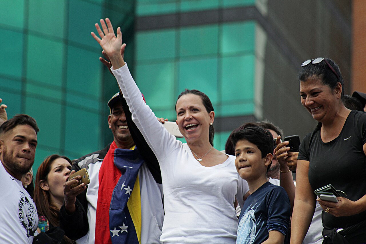 María Corina Machado, in a white top, smiles and waves to a crowd of supporters, one draped in a Venezuelan flag