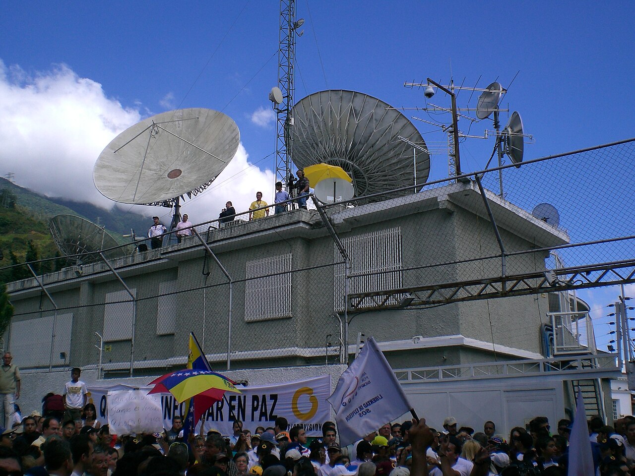 Satellite dishes and antennas on the roof of a grey broadcast building in Caracas, with a crowd holding flags and a banner below.