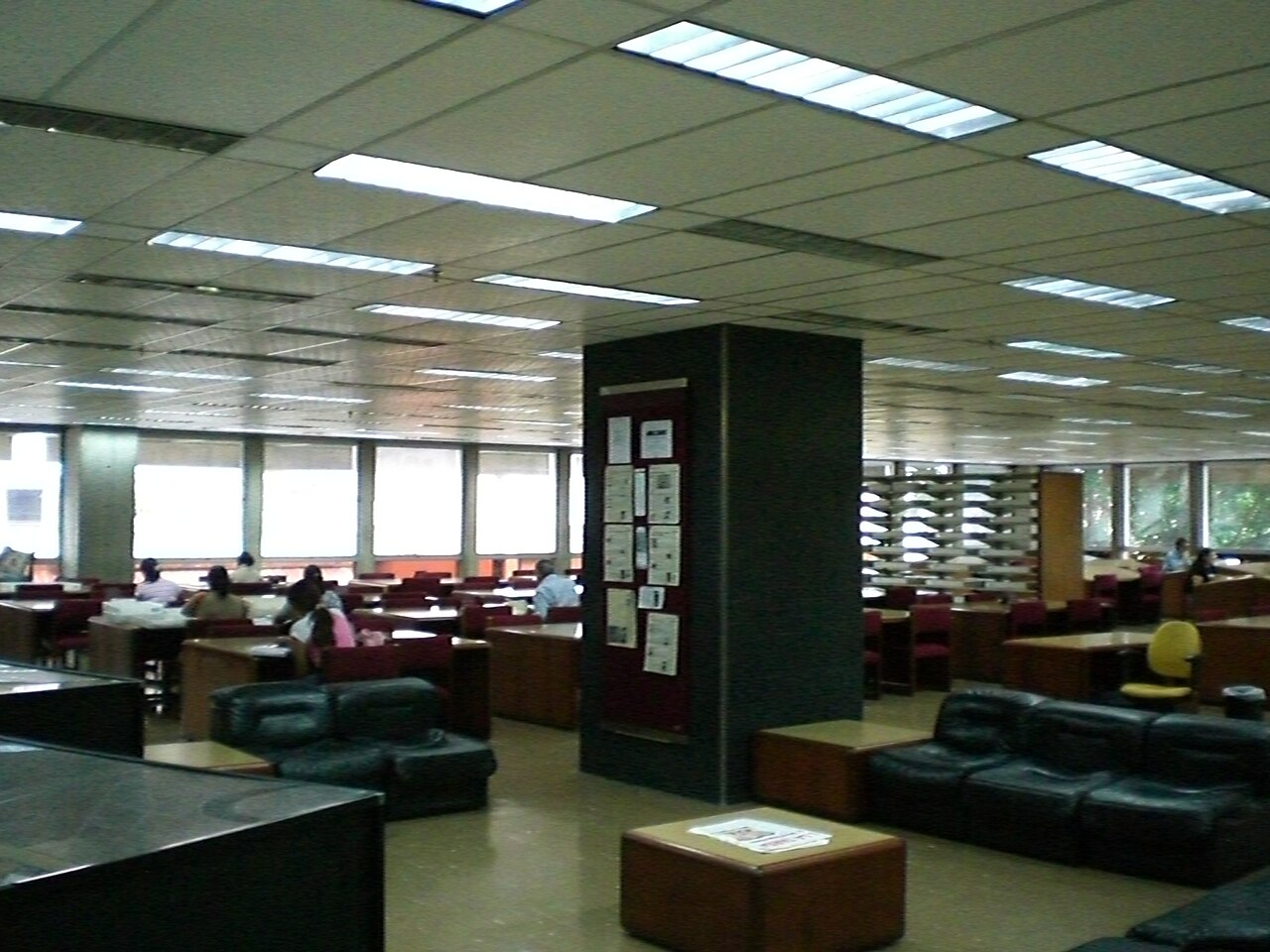 A large reading room with desks, black sofas and newspaper pages pinned to a central column in the National Library in Caracas.