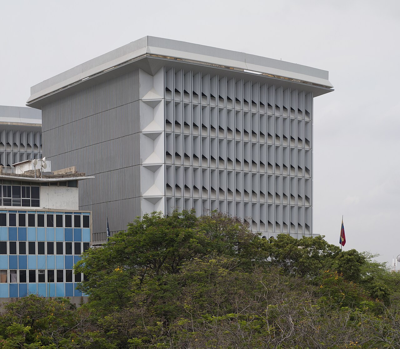 The grey concrete tower of the Central Bank of Venezuela in Maracaibo, with a Venezuelan flag on a pole and green trees in front