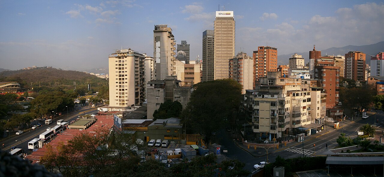 Apartment and office towers in central Caracas under a blue sky, with buses parked on a busy avenue and green hills behind