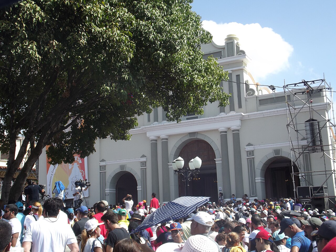 A large crowd of pilgrims in front of the cream-coloured Basilica of Santa Rosa in Barquisimeto, with a camera crew and a sound stage at the edge.