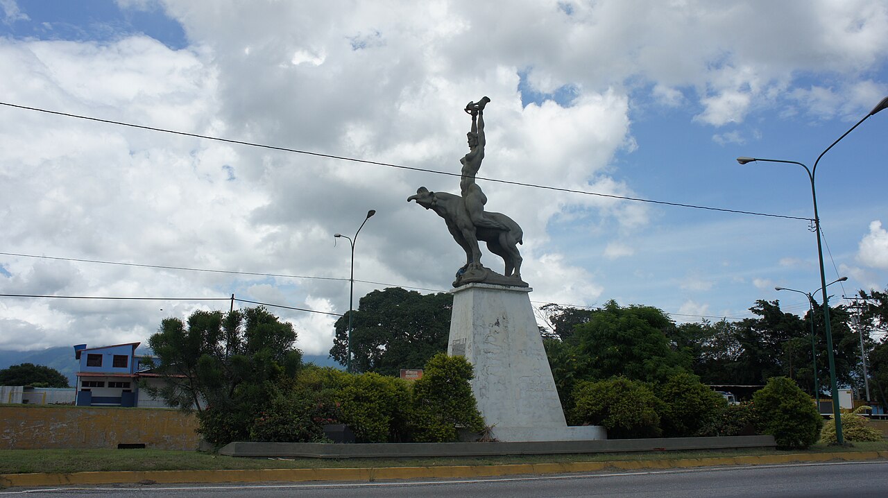 A stone statue of a woman raising a small object above her head and riding a large animal, on a white pedestal beside a road with green hills behind.