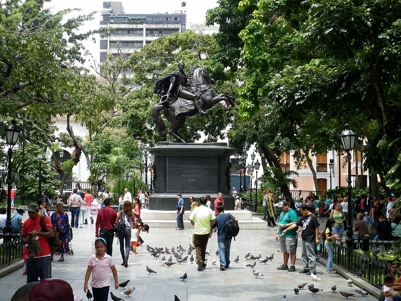 People and pigeons around the bronze equestrian statue of Simón Bolívar in Plaza Bolívar, central Caracas, under shady trees