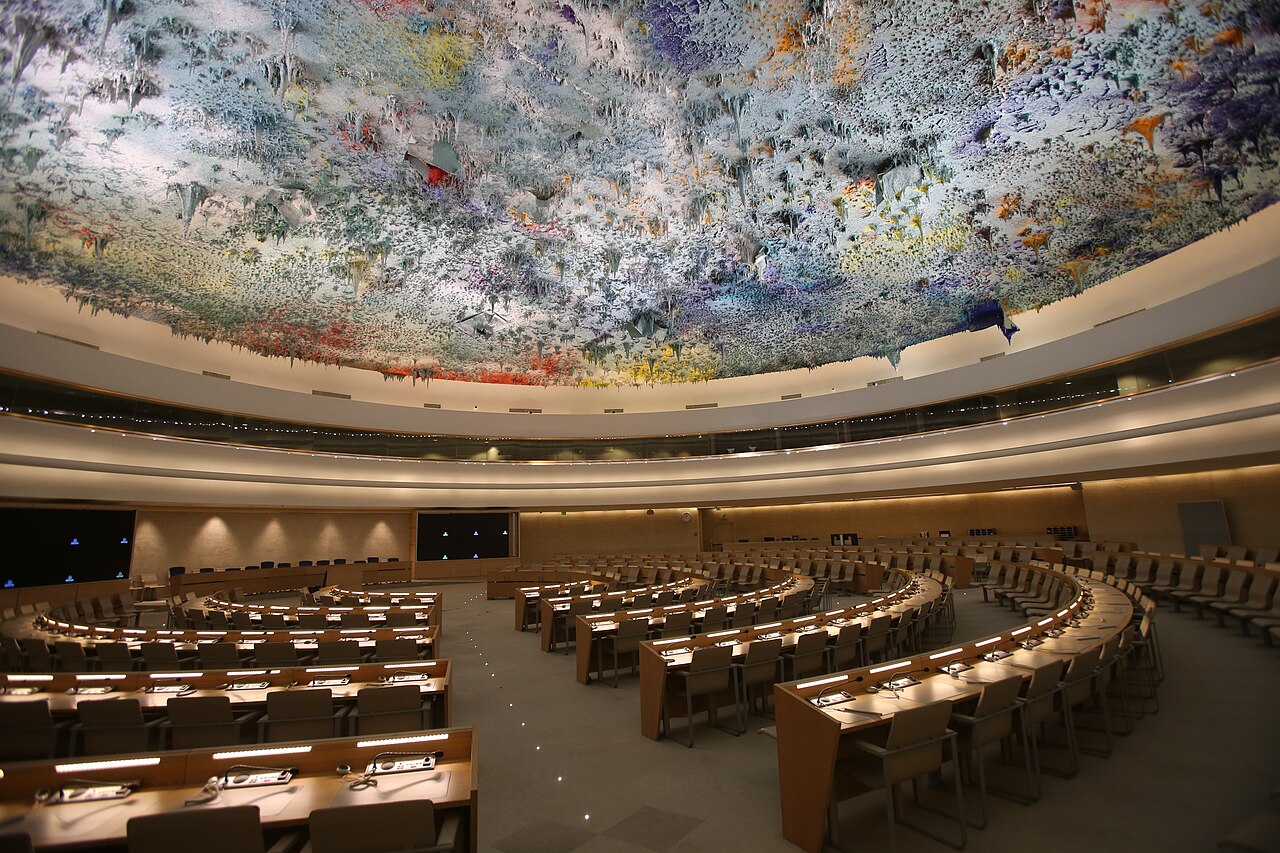 The Human Rights Council chamber at the Palais des Nations in Geneva, with curved rows of empty delegate desks under a colourful dripping-paint ceiling