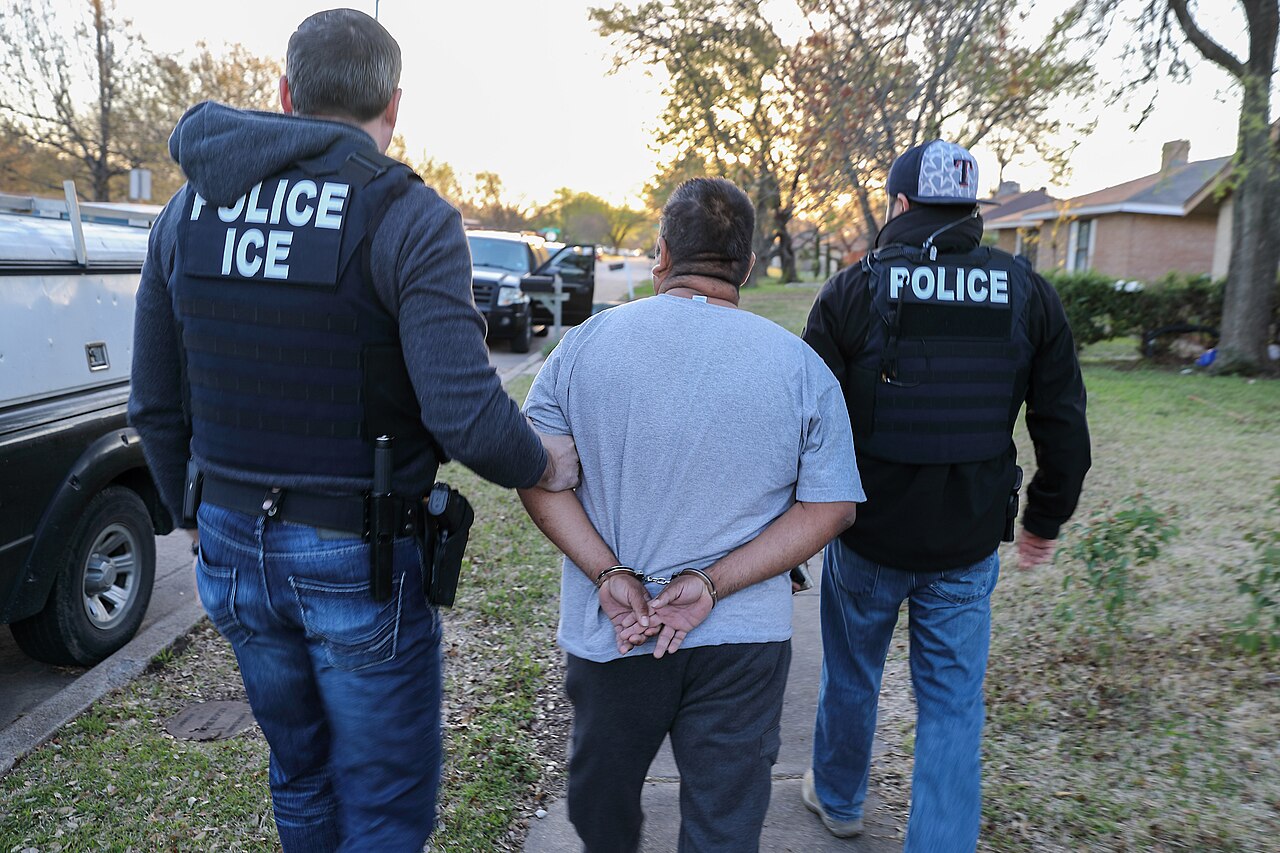 Two ICE officers in police vests walk a handcuffed man along a residential sidewalk in Texas