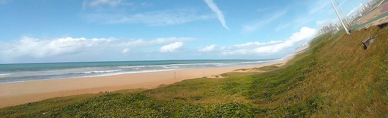 Wide view of a sandy Atlantic beach with waves, a grassy dune slope and city buildings along the seafront in the distance, in the Boca do Rio district of Salvador.