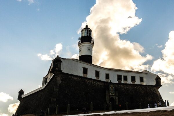 Black-and-white Farol da Barra lighthouse rising above the stone walls of the Santo Antônio da Barra fort in Salvador under a blue sky with large clouds.