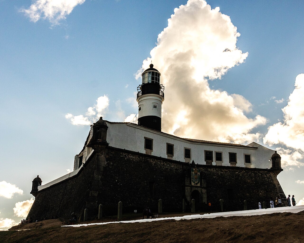 Black-and-white Farol da Barra lighthouse rising above the stone walls of the Santo Antônio da Barra fort in Salvador under a blue sky with large clouds.