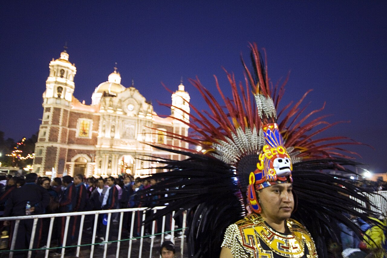 A dancer in a feathered headdress stands outside the floodlit Old Basilica of Guadalupe at night while pilgrims queue behind a barrier