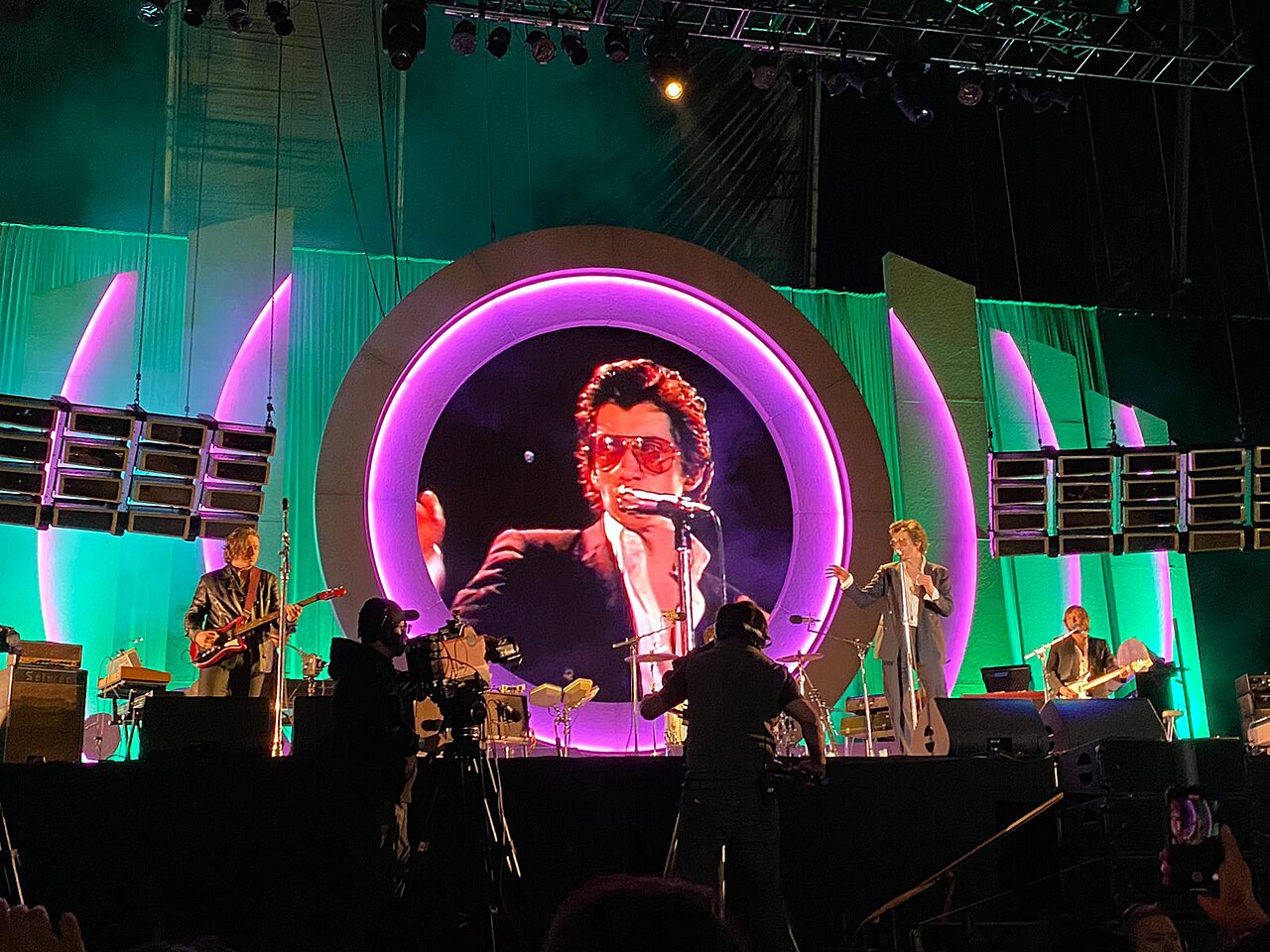 Rock band on a stage lit in green and purple, with a large circular screen showing the singer in sunglasses, at a stadium concert in Mexico City