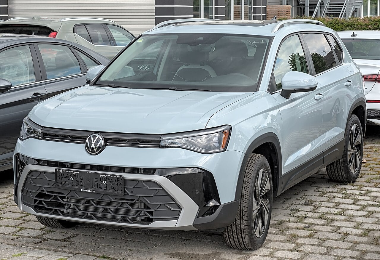 A light-blue Volkswagen Taos compact SUV parked on a cobbled lot in front of a car dealership