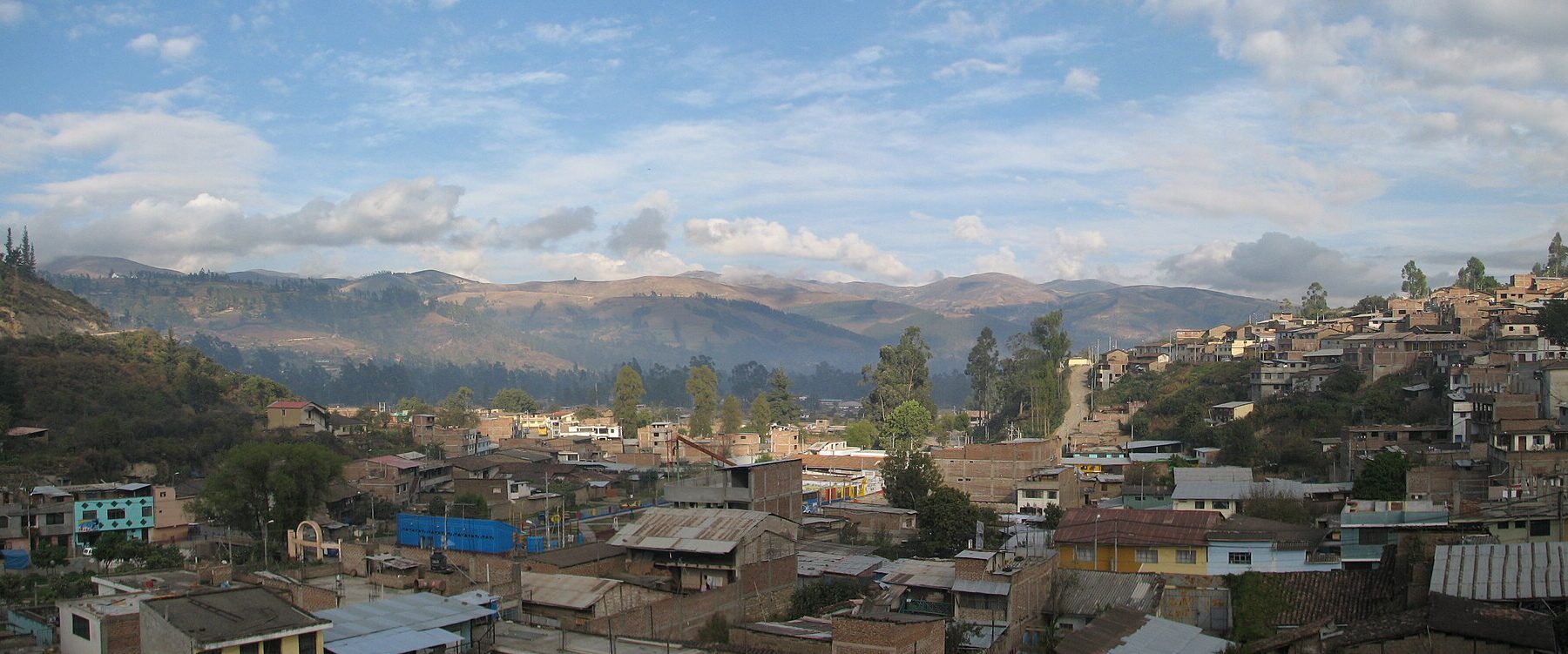 Panoramic view over a hillside Andean town of brick and tin-roofed houses, with eucalyptus trees and brown mountains under a blue sky with clouds