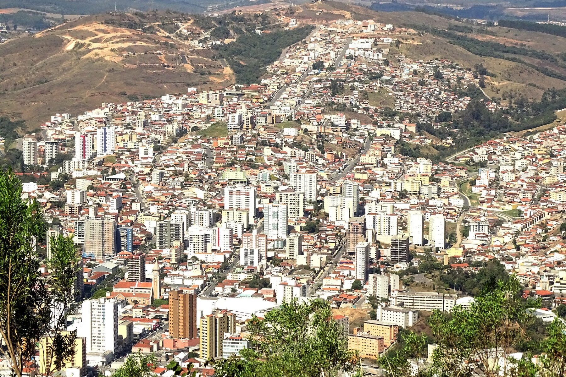Elevated view of Poços de Caldas, a hillside city of white high-rise and low red-roofed buildings, with dry hills and bare earth slopes behind it
