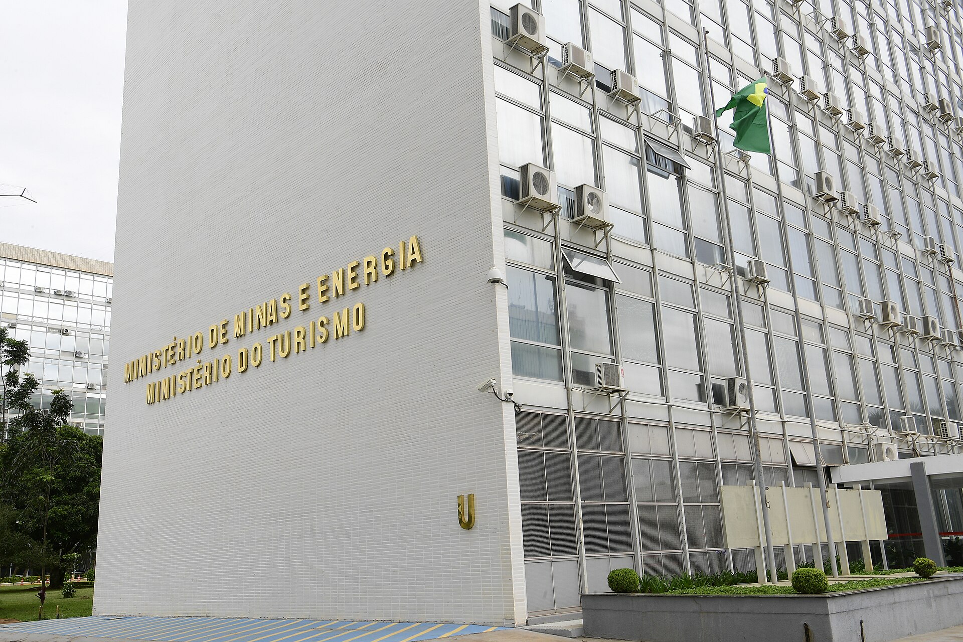 A white modernist office block in Brasília with gold lettering reading Ministério de Minas e Energia and Ministério do Turismo, a Brazilian flag on the facade