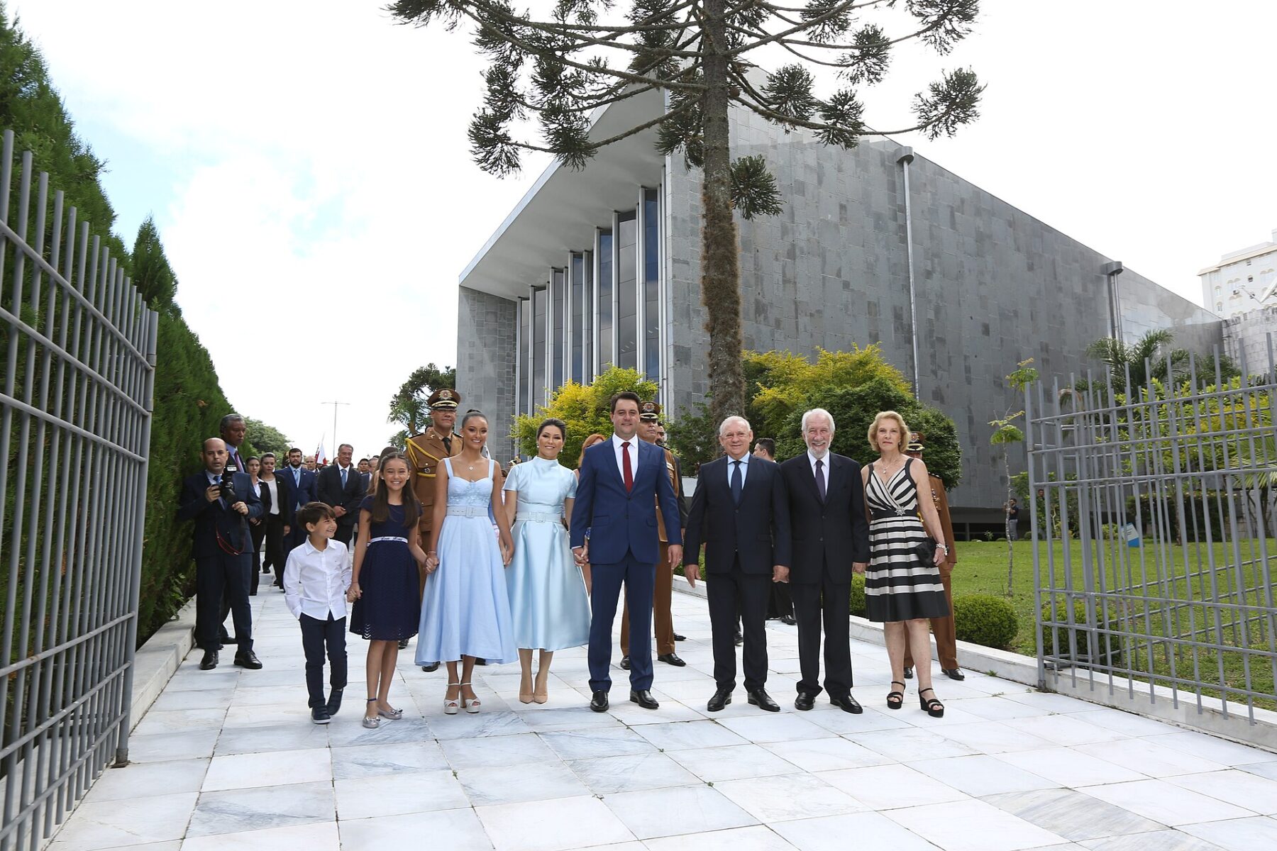 A group in formal clothes, including children, poses on a white stone path in front of a large grey modernist building with an araucaria tree
