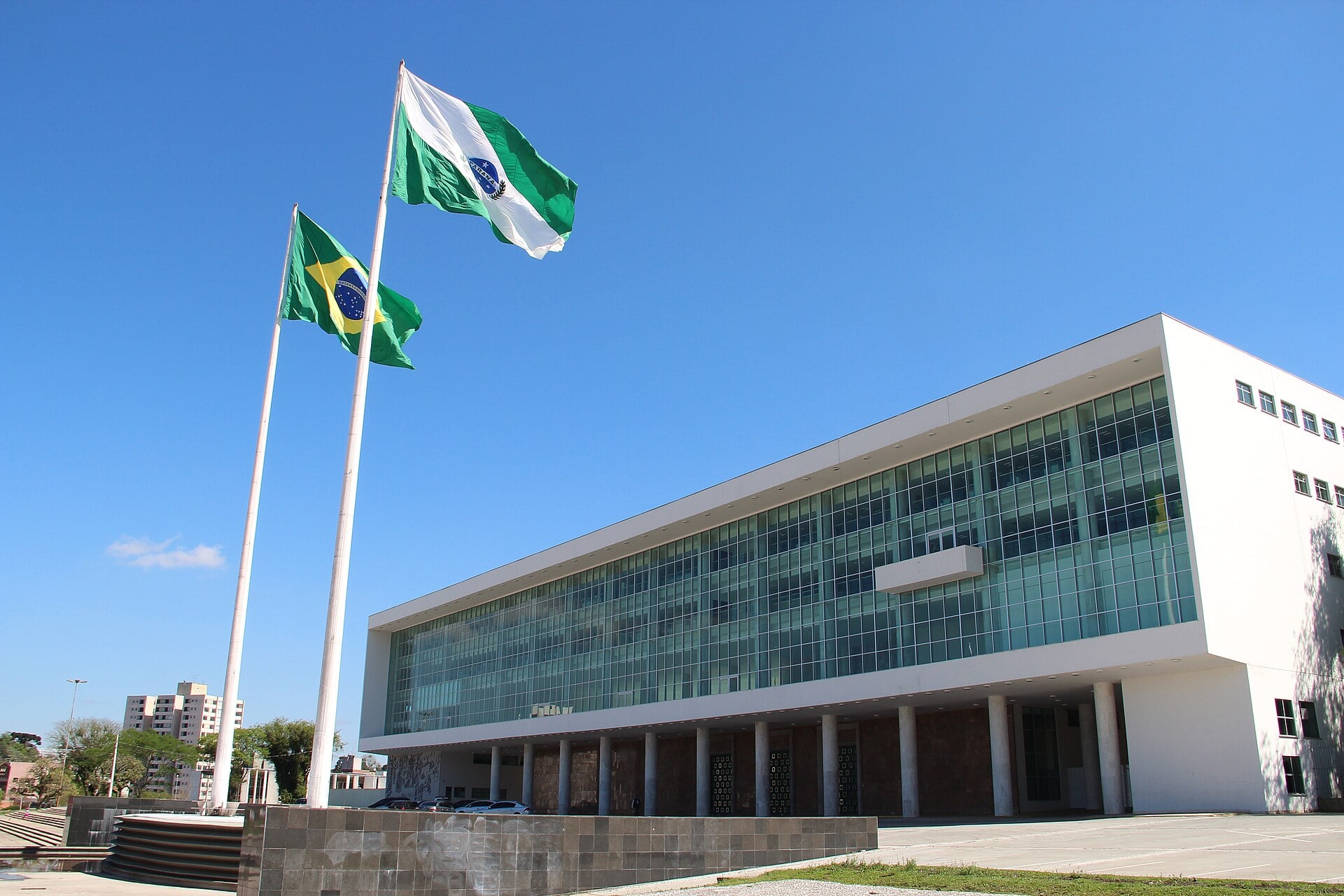 A long modernist glass-and-white office building under a blue sky, with the Brazilian flag and the green-and-white Paraná state flag on tall poles