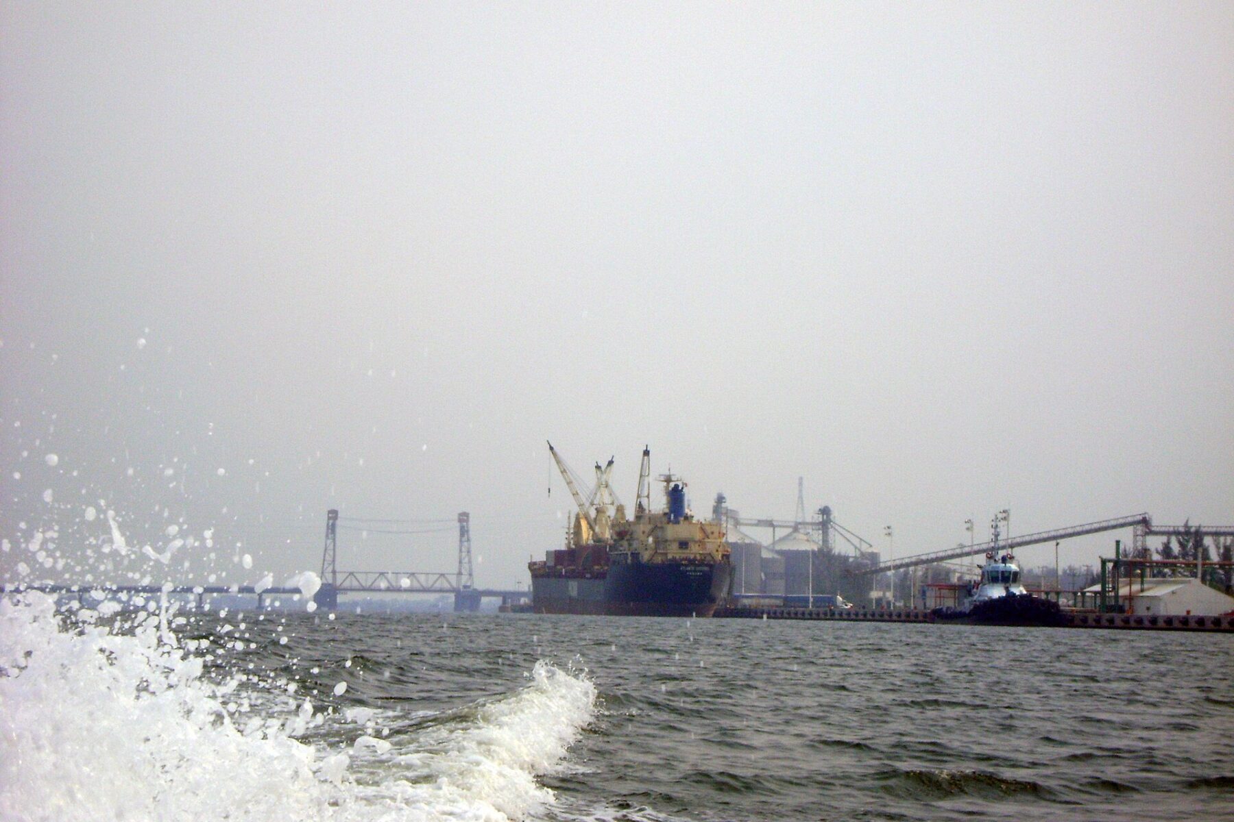 A cargo ship and a tugboat moored at an industrial river port with cranes and conveyor belts, a lift bridge in the hazy background and spray in the foreground