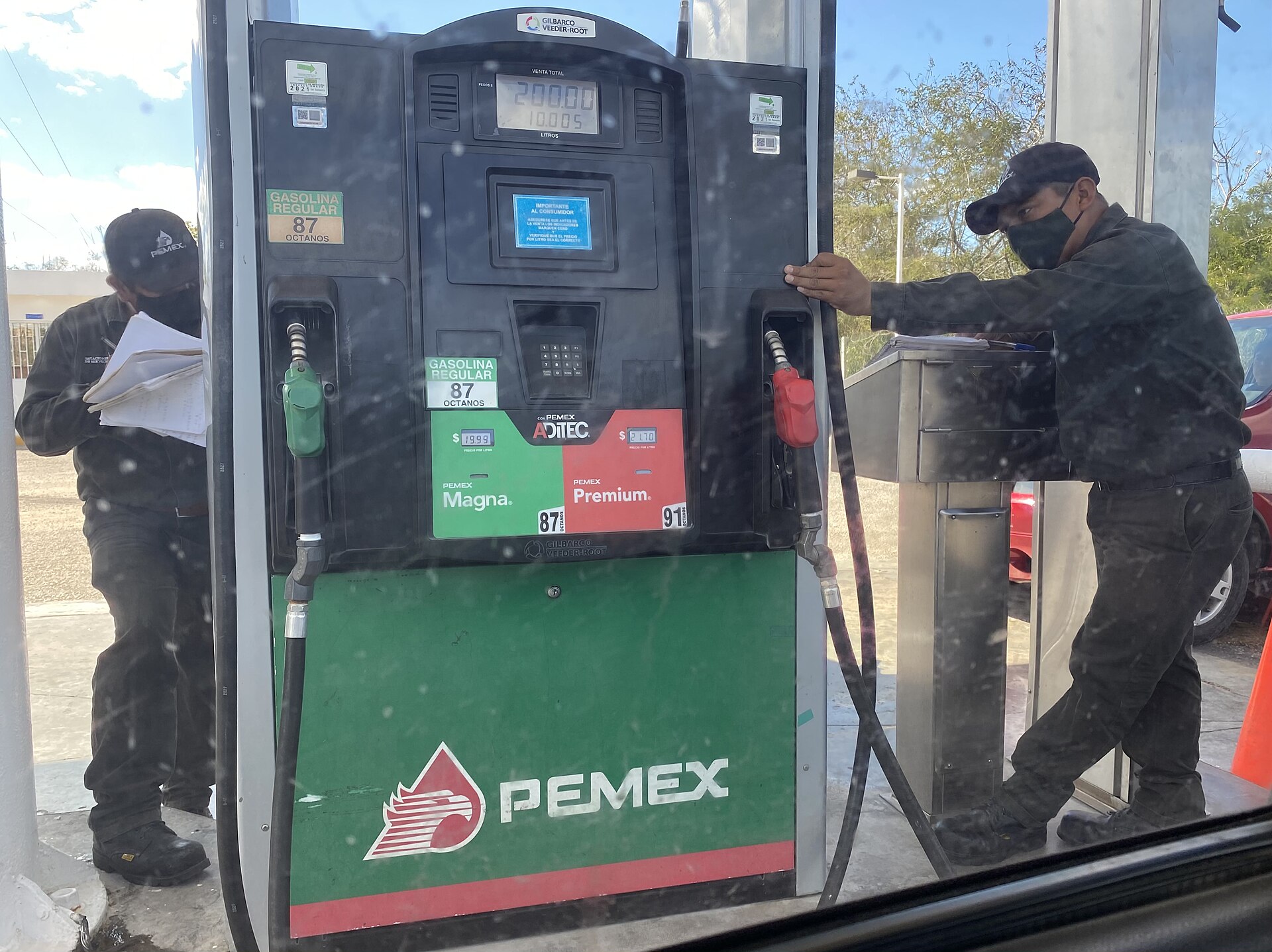 Two uniformed attendants beside a green and red Pemex fuel dispenser showing Magna and Premium gasoline, photographed through a car window