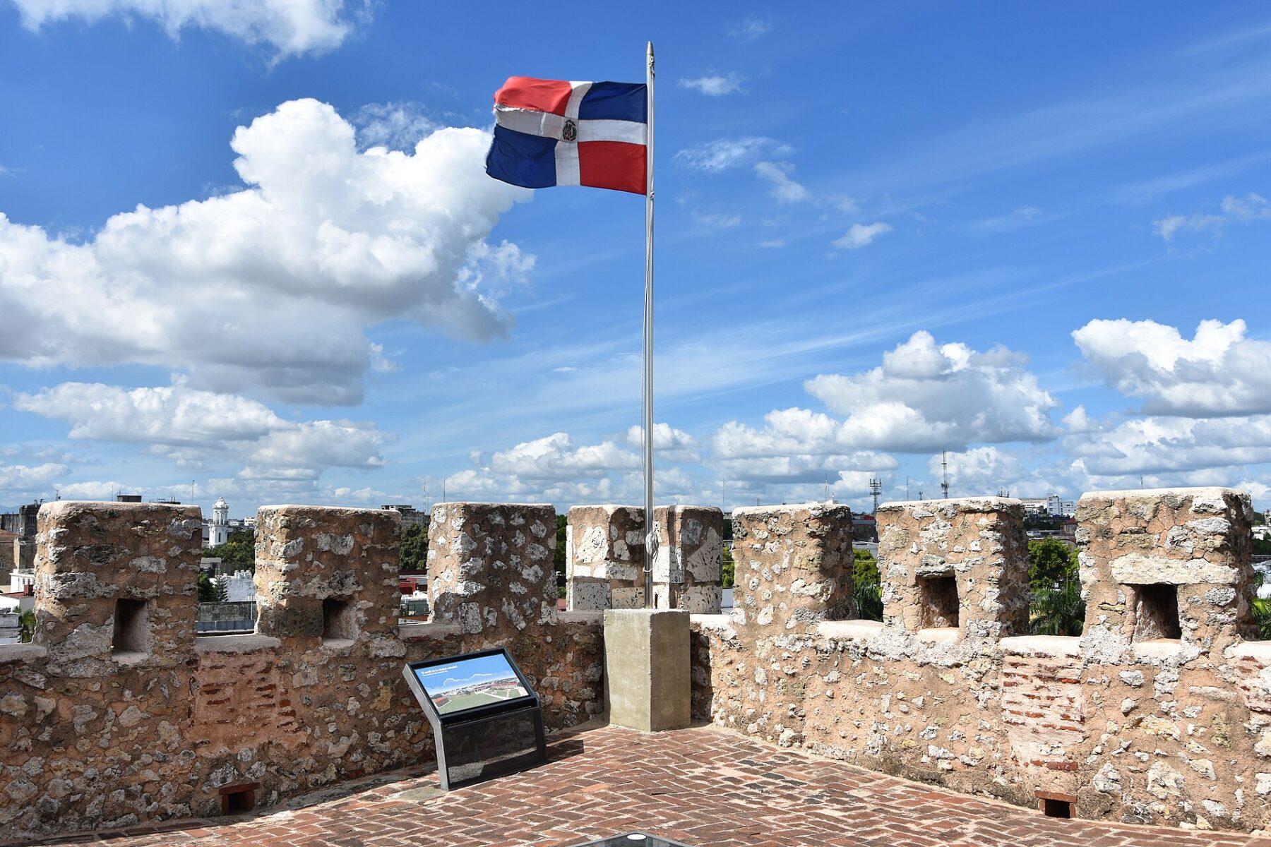 The Dominican flag flying on a pole above the stone battlements of a colonial fortress under a blue sky with white clouds