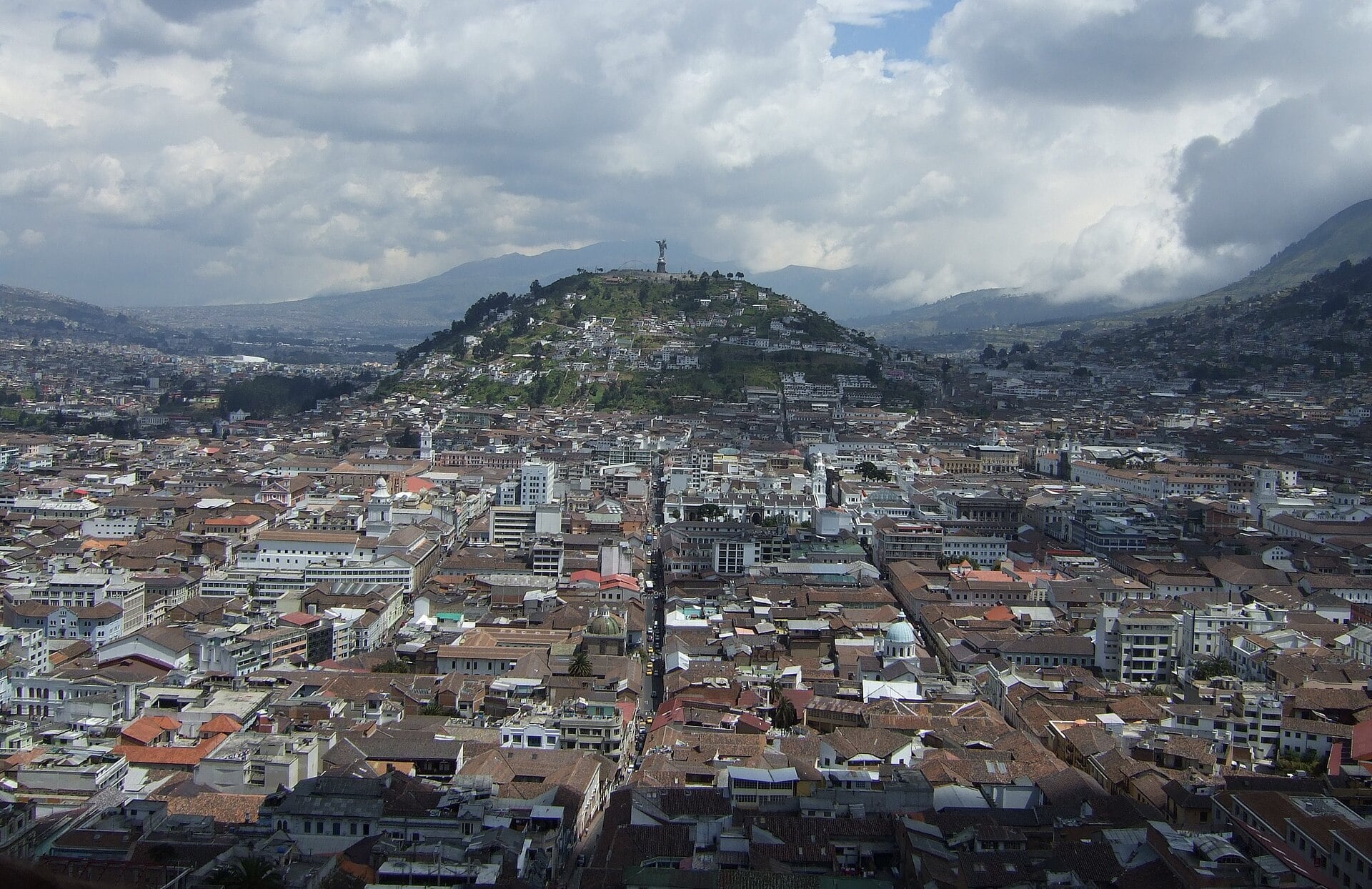 Dense rooftops of central Quito stretching towards a green hill topped by a winged Virgin statue, with Andean mountains and clouds behind