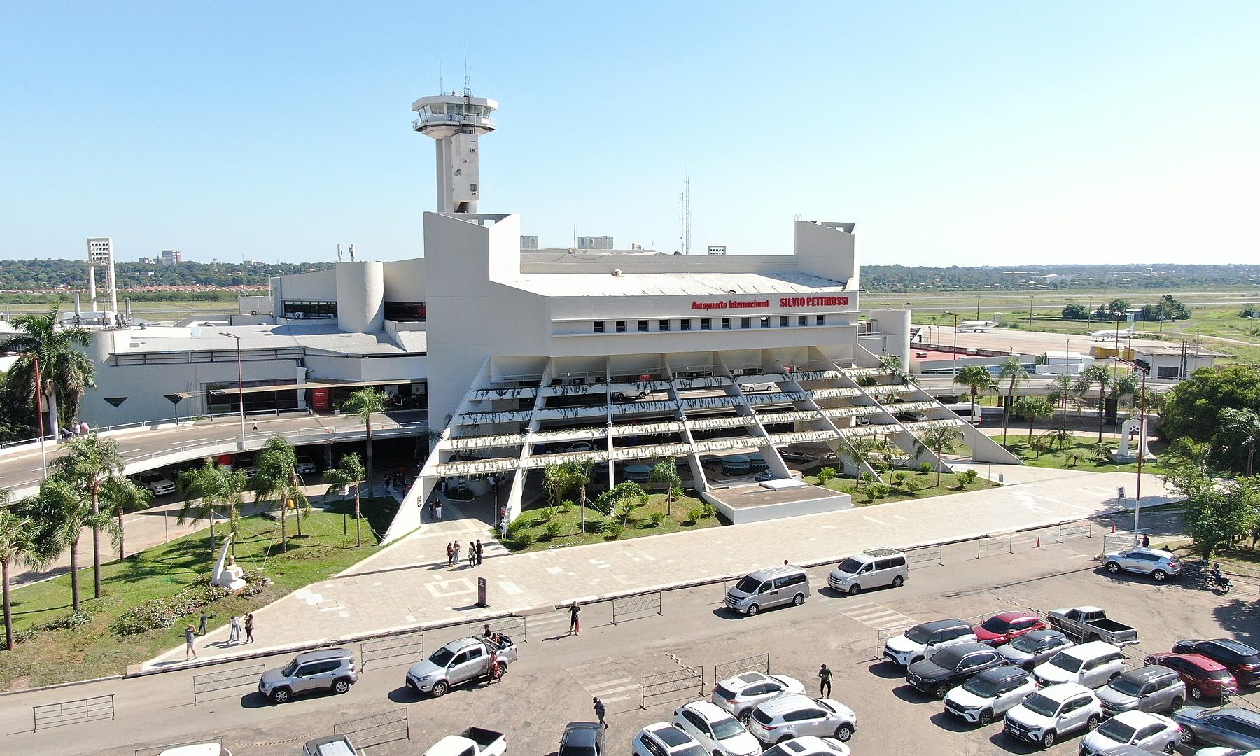 Aerial view of a white airport terminal with a control tower, palm trees, a pedestrian plaza and a car park under a clear blue sky