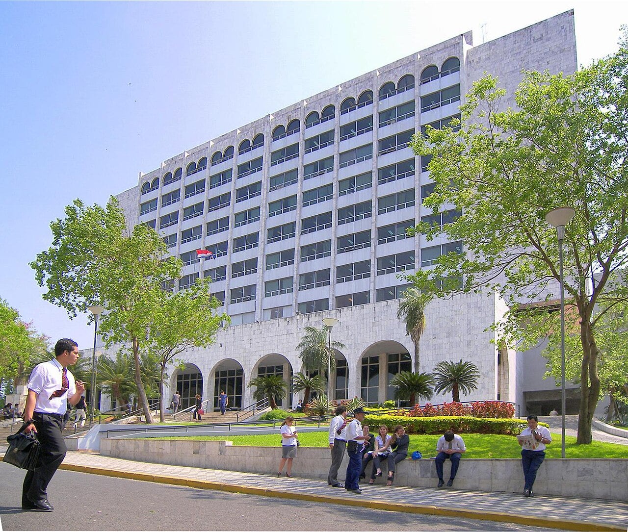 A tall white stone court building with arched windows on the top floor, trees in front and people walking and sitting on a low wall