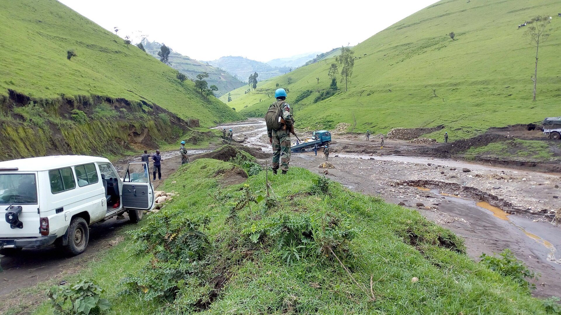A UN peacekeeper in a blue helmet overlooks a muddy, damaged stretch of road in a green valley in Masisi territory