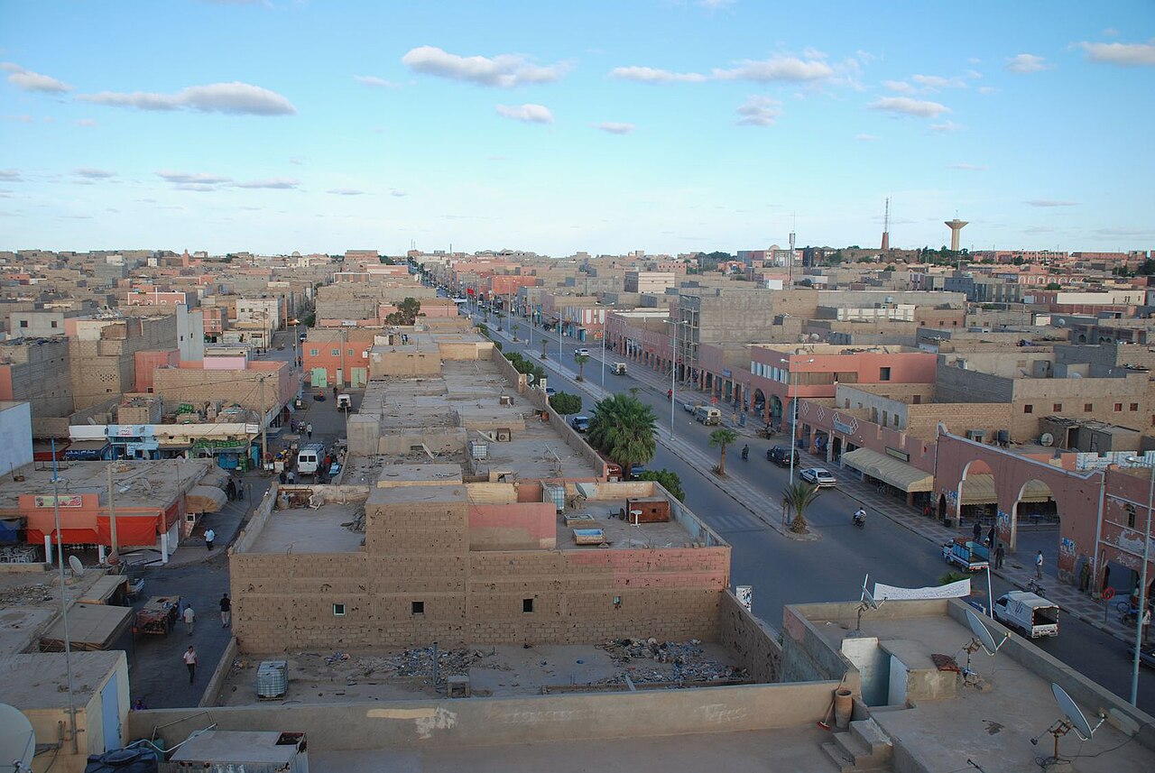 Main road through the town of Smara in Western Sahara, seen from a rooftop, with low buildings, palm trees and a water tower under a blue sky
