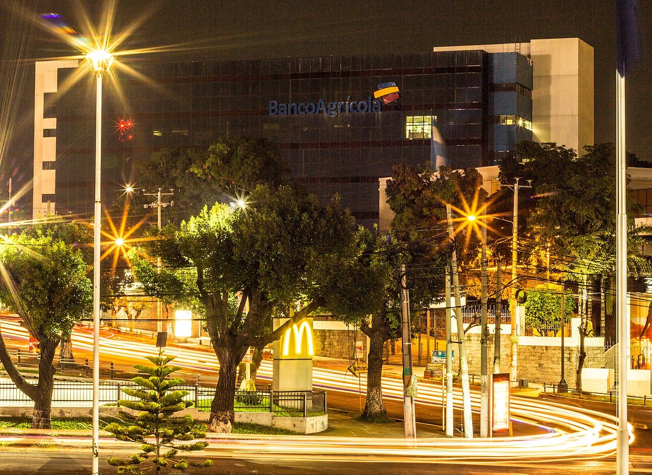 A Banco Agrícola office building with the bank's name and logo, seen at night behind trees and light trails from traffic
